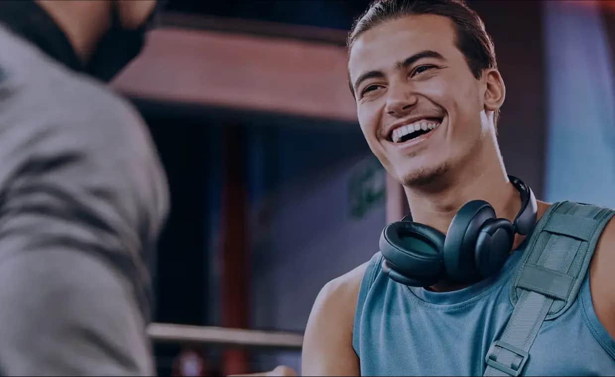 A young man wearing headphones around his neck and a tank top smiles while talking to another person at a gym. The atmosphere is friendly and casual.