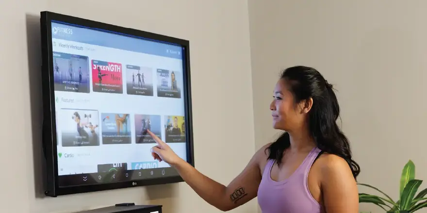 A woman in a purple tank top touches a wall-mounted touchscreen displaying various workout videos, selecting an option.
