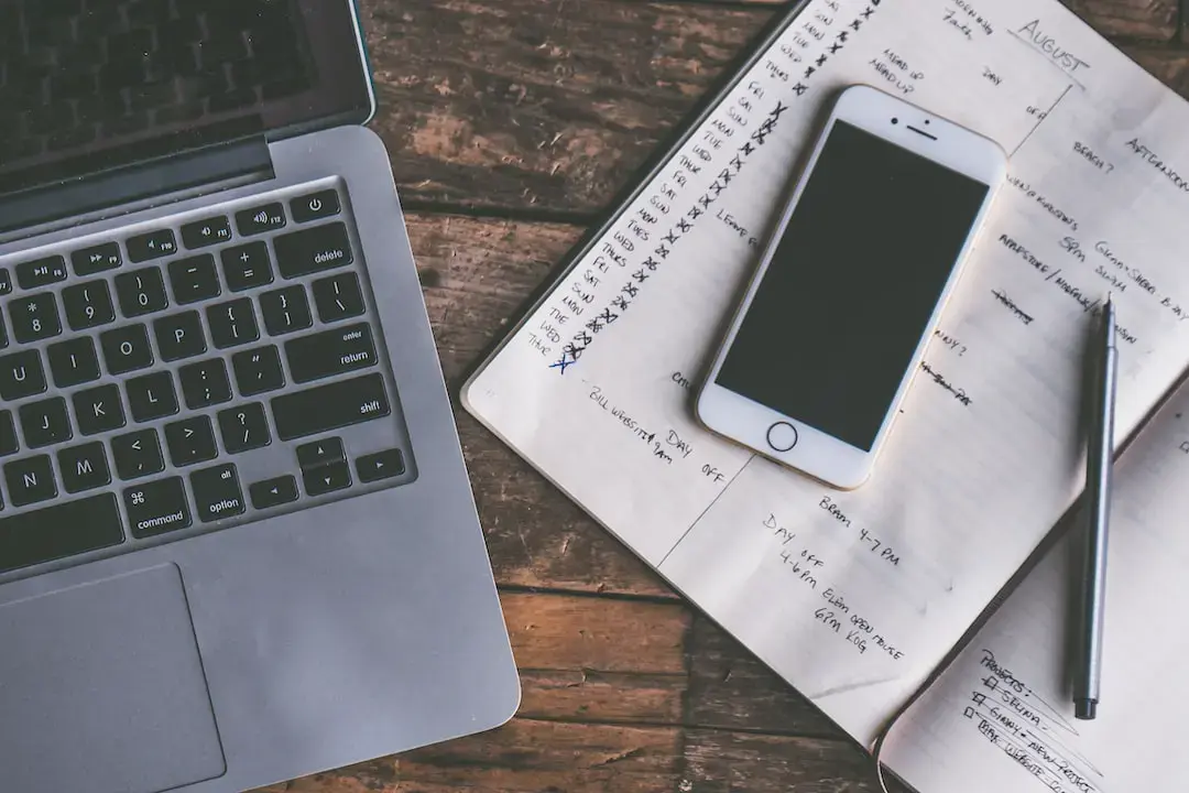 A laptop, smartphone, and pen rest on an open planner with handwritten notes and dates for gym schedules, all placed on a rustic wooden surface.