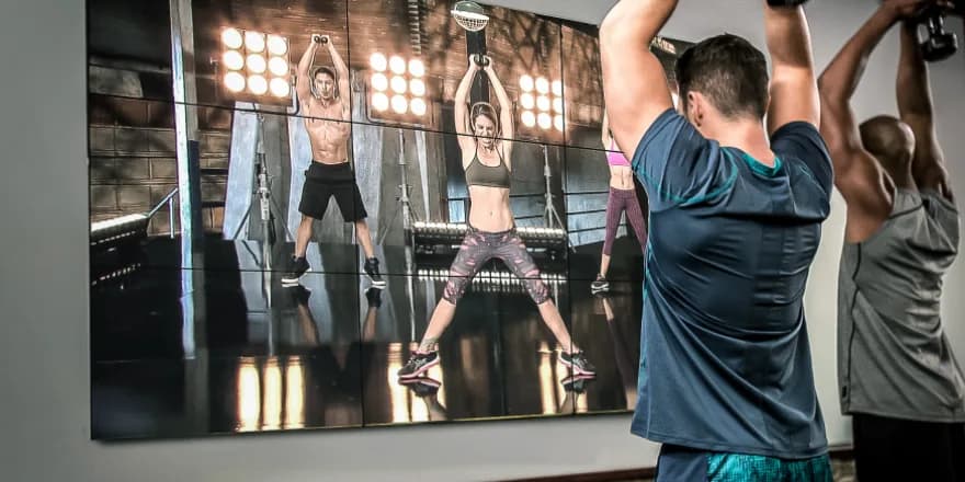 Two men in athletic wear exercise with dumbbells in front of a large screen showing a workout video featuring three instructors lifting weights in a studio with bright lights.