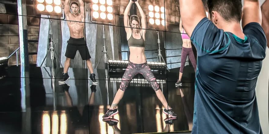 Two people in a gym follow a workout video on a large screen, which shows three fitness instructors performing aerobic exercises in athletic wear on a brightly lit stage.