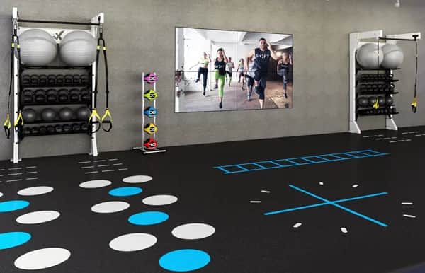 A modern gym room with exercise balls, kettlebells, weight racks, and resistance bands on both sides. The floor has blue and white workout markings, and a large screen displays a fitness class in progress.
