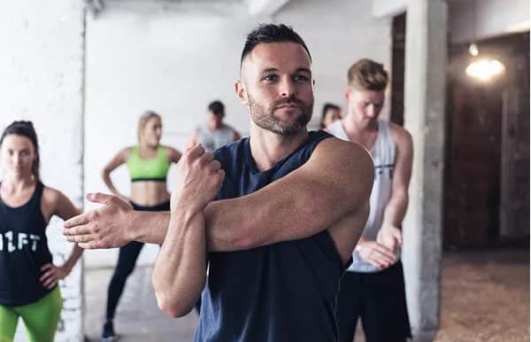A man in a sleeveless shirt stretches his arm across his chest in a gym, with four other people in athletic wear stretching in the background.
