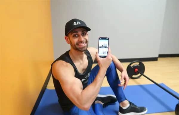 A man in athletic wear sits on a blue exercise mat in a gym, smiling and holding up a smartphone displaying an image. A barbell is on the floor beside him, and the background features yellow and gray walls.