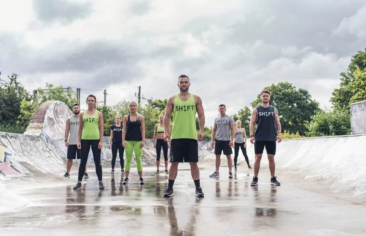 A group of people wearing SHIFT workout clothes stand in formation on a wet outdoor skatepark, with greenery and cloudy skies in the background.