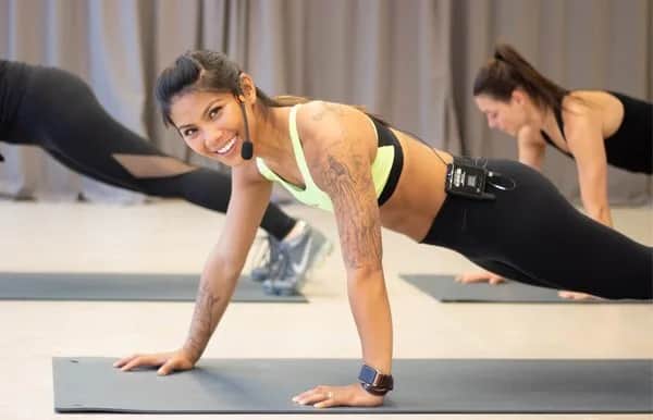 A smiling fitness instructor with tattoos leads a workout class, performing a plank on a yoga mat while wearing a headset microphone and athletic clothing. Other participants are also planking in the background.