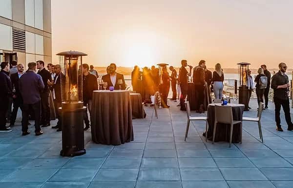 People gather on a rooftop terrace at sunset, standing and conversing around tall tables with heaters. Some people are seated, while others mingle, enjoying the view and ambiance.