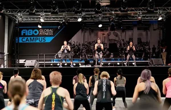 A group fitness class takes place at FIBO Global Fitness Campus, with three instructors leading exercises on a stage in front of an audience wearing athletic clothing.