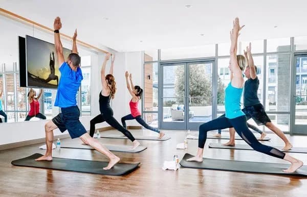Four people practice yoga in a bright studio, following an instructor on a screen. They are in warrior pose on black mats, with water bottles and towels nearby. Large windows let in natural light.