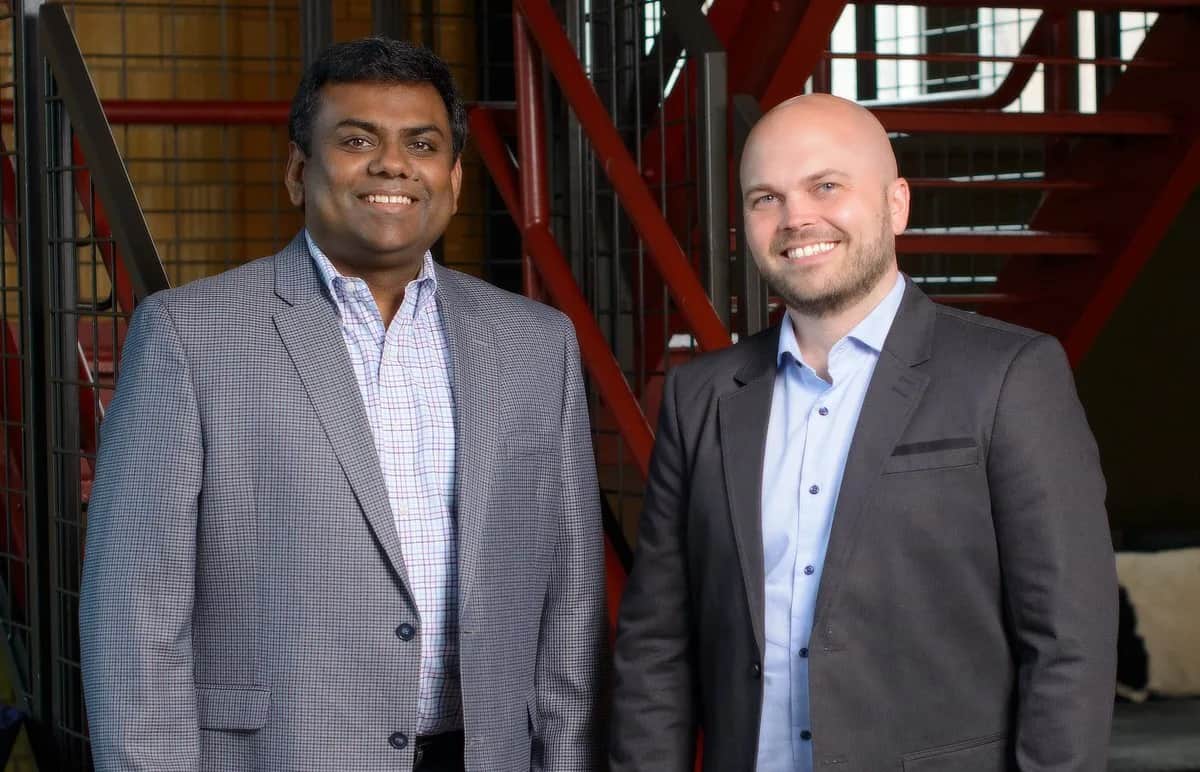 Two men in business attire stand side by side, smiling at the camera. They are in an indoor setting with metal railings and a red staircase in the background.