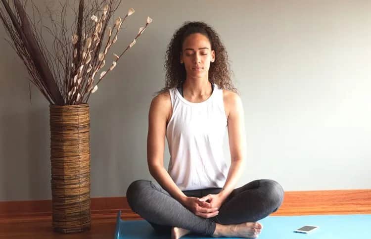 A woman sits cross-legged on a yoga mat indoors with eyes closed, hands resting on her lap, near a tall vase of decorative branches and a smartphone on the floor beside her.