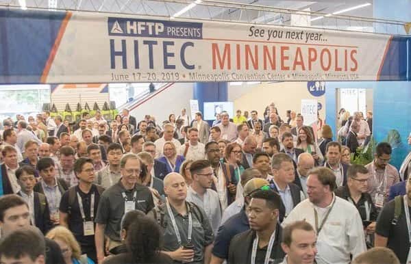 A large crowd of people attending a conference walk under a banner that reads “HITEC MINNEAPOLIS, June 17-20, 2019, Minneapolis Convention Center.” The scene is busy with attendees wearing badges.
