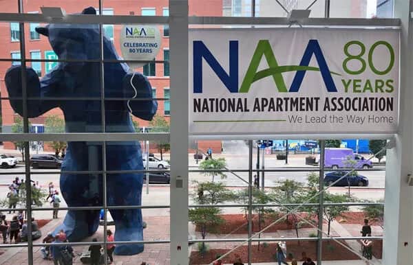 A large blue bear sculpture peers into a glass building. Inside, a banner reads NAA 80 Years National Apartment Association, We Lead the Way Home. People walk on the sidewalk outside.