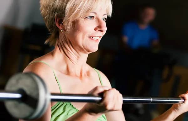 A smiling woman with short blonde hair lifts a barbell indoors, wearing a green sleeveless top. The background is blurred, with another person faintly visible.