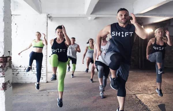 A group fitness class is exercising indoors, with participants wearing athletic clothing and performing high knee movements in unison. The instructor leads at the front, and the room has white brick walls and lots of natural light.