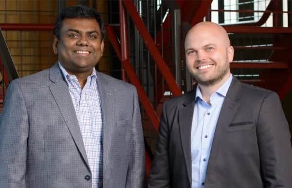 Two men in business attire stand side by side, smiling at the camera. One has dark hair and is wearing a gray blazer and checked shirt, while the other is bald and wearing a dark blazer and white shirt. Red stairs are in the background.
