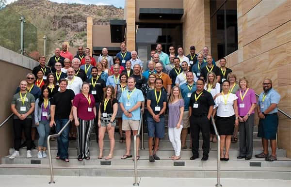 A large group of people wearing name tags and yellow lanyards stands and smiles on outdoor steps in front of a modern building, with rocky hills and greenery visible in the background.