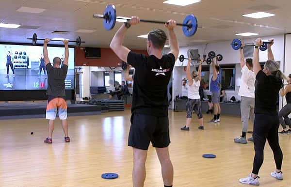 A group fitness class lifts barbells overhead in a gym with wood floors, mirrors, and a large screen displaying workout instructions. Participants are spaced out and following along with the virtual instructor.