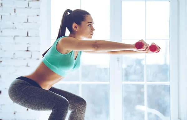 A woman in athletic wear performs a squat indoors, holding small red dumbbells and extending her arms forward. She appears focused, with light streaming in from large windows behind her.