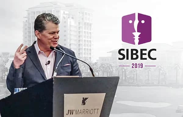 A man in a suit speaks at a podium labeled JW Marriott with the SIBEC 2019 logo displayed beside him; a building and palm trees are visible in the background.