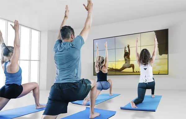 Four people on yoga mats follow a virtual yoga instructor displayed on a large screen in a bright, modern room. They are all performing a lunge pose with arms raised, facing the screen.