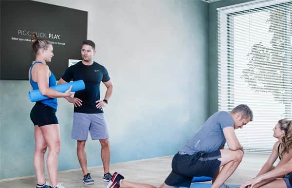 Four people are in a bright fitness studio with wooden floors. Two are standing and talking, holding yoga mats, while two others are stretching on the floor. A large screen on the wall displays workout instructions.