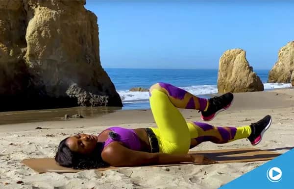 A woman in bright workout clothes lies on a mat at the beach, performing a leg exercise with bent knees. Large rocks and the ocean are visible in the background under a clear blue sky.