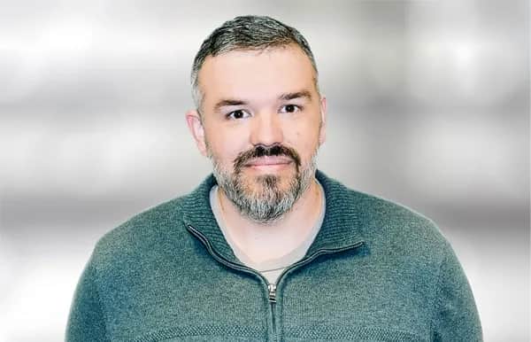A man with short dark hair and a beard is wearing a greenish-gray zip-up sweater. He is looking at the camera and standing against a blurred light gray background.