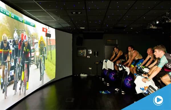 A group of cyclists on stationary bikes rides indoors, watching a large screen showing a professional cycling race. The room is dark with ceiling lights, and the cyclists are focused on the race.