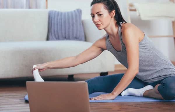 A woman in workout clothes sits on a yoga mat at home, stretching toward her foot while looking at a laptop screen in front of her. A sofa and blankets are visible in the background.