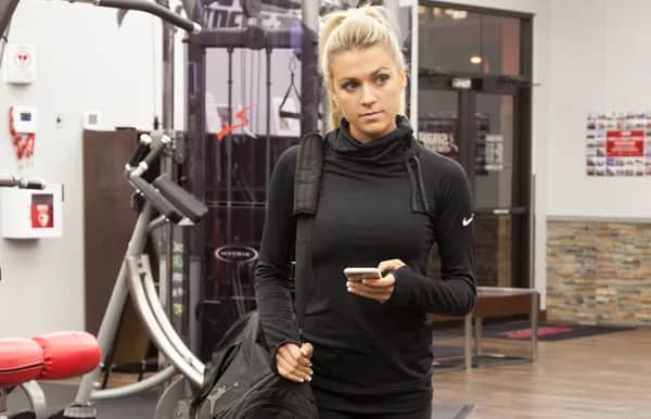 A woman in athletic wear stands in a gym holding a duffel bag and a smartphone, looking off to the side. Gym equipment and a bench are visible in the background.