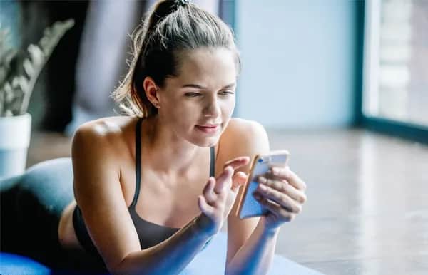 A woman in workout clothes lies on a yoga mat indoors, looking at her smartphone and smiling slightly. Natural light comes through large windows in the background.
