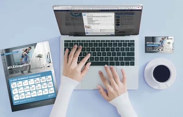 A person types on a laptop at a desk with a fitness challenge flyer, a coffee cup, and two cards nearby, all on a light blue surface.