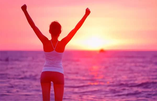 A woman stands by the ocean with her arms raised in triumph, facing a vibrant pink and orange sunset. She wears a tank top and shorts, celebrating or enjoying the serene beach view.
