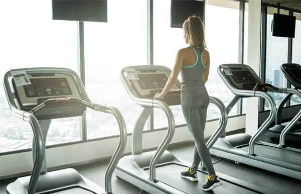 A woman in athletic wear stands on a treadmill in a bright, modern gym with large windows and several other treadmills nearby.