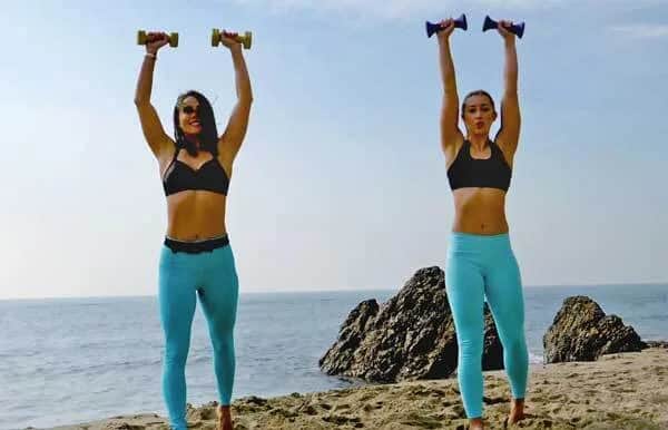 Two women wearing black sports bras and blue leggings exercise with dumbbells on a sandy beach near the ocean, with large rocks in the background. Both are raising dumbbells above their heads under a clear sky.