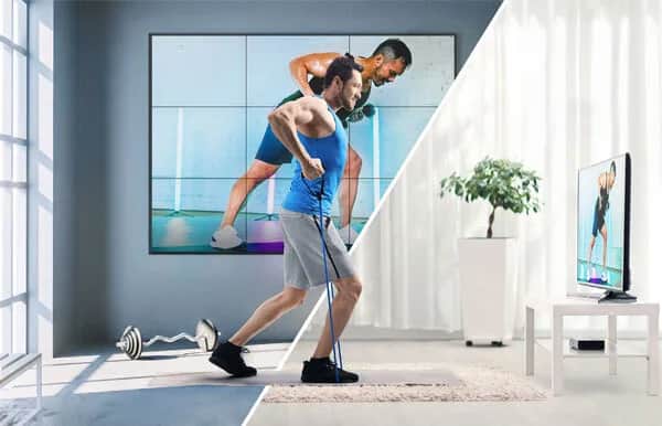 A man exercises at home with resistance bands, following a workout on a large screen. The image is split: one side shows a bright, modern gym; the other, a cozy living room with a TV displaying the same workout.