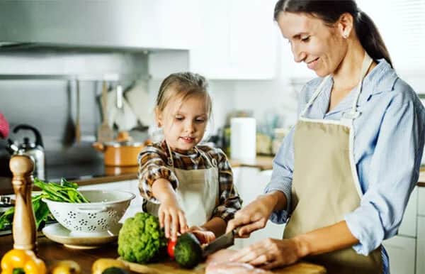A woman and a young girl wearing aprons prepare food together in a bright kitchen. The woman cuts meat while the girl reaches for fresh vegetables on the counter. They both look focused and happy.