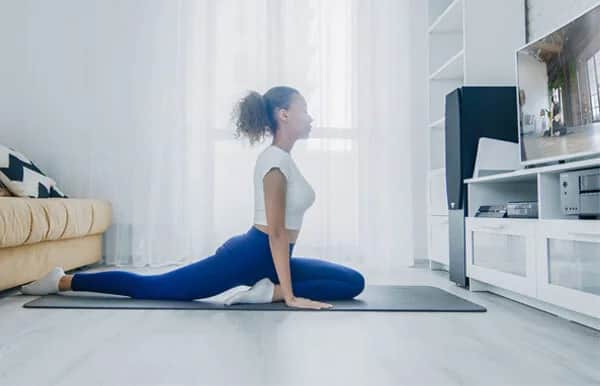 A woman in athletic wear practices yoga on a mat in her living room, following a routine displayed on a TV. She is doing a pigeon pose, with bright natural light coming through sheer curtains behind her.
