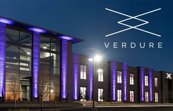 Modern building with large windows lit by purple lights at night. The word “Verdure” and its logo appear in the sky above the building. The area in front of the building has young trees and a sidewalk.