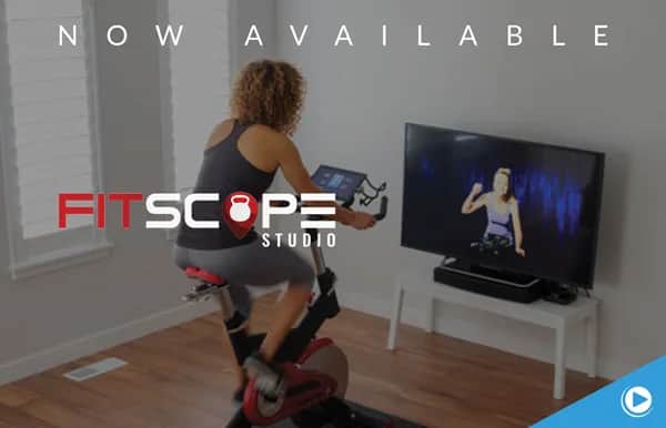 A woman rides an exercise bike indoors, following a virtual fitness class on a TV. Text reads Now Available and Fitscope Studio with a kettlebell icon. The room has wood floors and large windows.