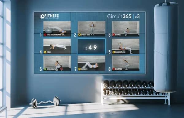 A gym room with weights and exercise balls, featuring a large digital screen displaying a workout circuit with images of eight different exercises and a timer counting down from 49 seconds.
