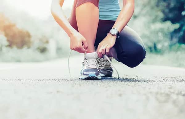 A person in athletic clothing is kneeling on an outdoor path, tying the laces of their running shoes. The background is blurred with greenery, suggesting a park or nature trail.
