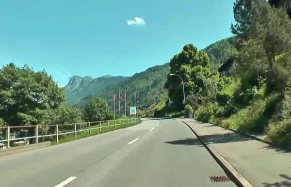 A two-lane road curves through a green, mountainous landscape under a bright blue sky. Trees line the road, and a few red flags are visible on the left side. One small cloud floats above the distant hills.