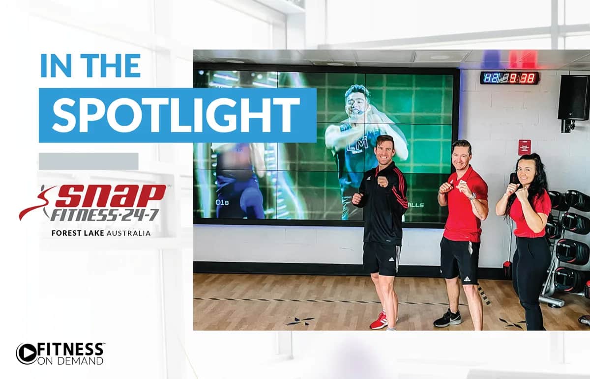 Three people posing in boxing stances in a gym, standing in front of a large screen displaying a male athlete. The text reads In the Spotlight and Snap Fitness 24-7 Forest Lake Australia. Fitness On Demand logo is also visible.