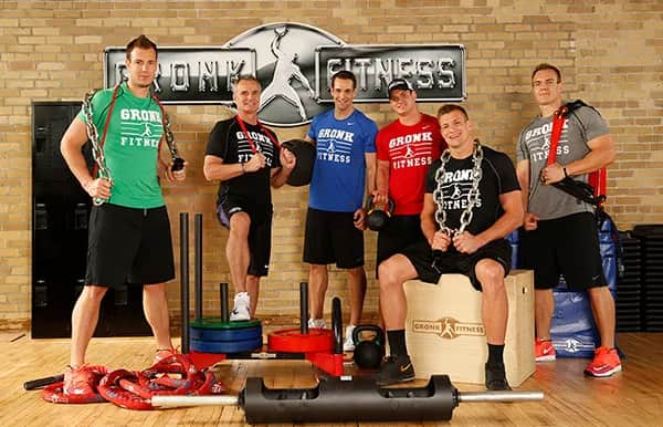 Six men in athletic wear pose in a gym with various fitness equipment, such as chains, ropes, and weights. Behind them is a Gronk Fitness sign on a brick wall. They are smiling and appear ready for a workout.