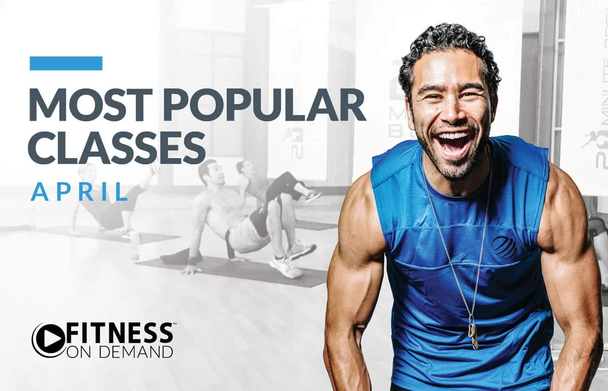A smiling, muscular man in a blue sleeveless shirt stands in the foreground; behind him, a group exercises in a fitness studio. Text reads Most Popular Classes April and Fitness On Demand.