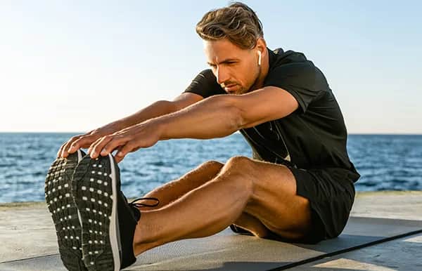 A man in athletic wear stretches forward to touch his toes while sitting on a mat near the ocean, with earbuds in his ears, during a sunny day.