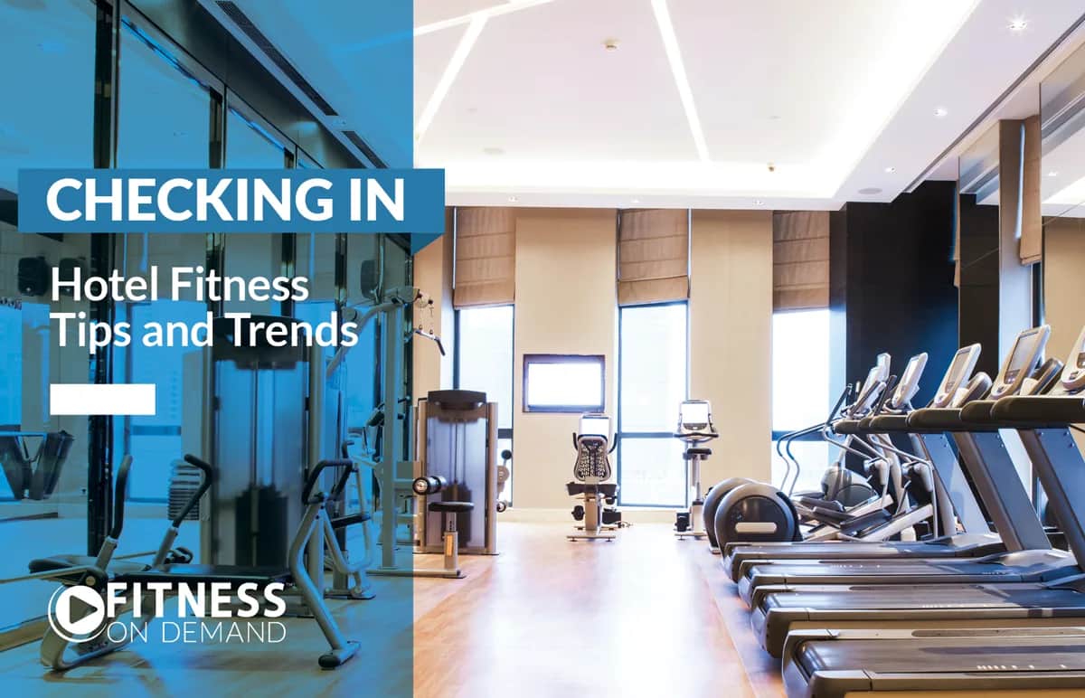 Modern hotel gym with treadmills, exercise bikes, and weight machines; large windows let in natural light. Text overlay reads: “Checking In: Hotel Fitness Tips and Trends. Fitness On Demand.”.
