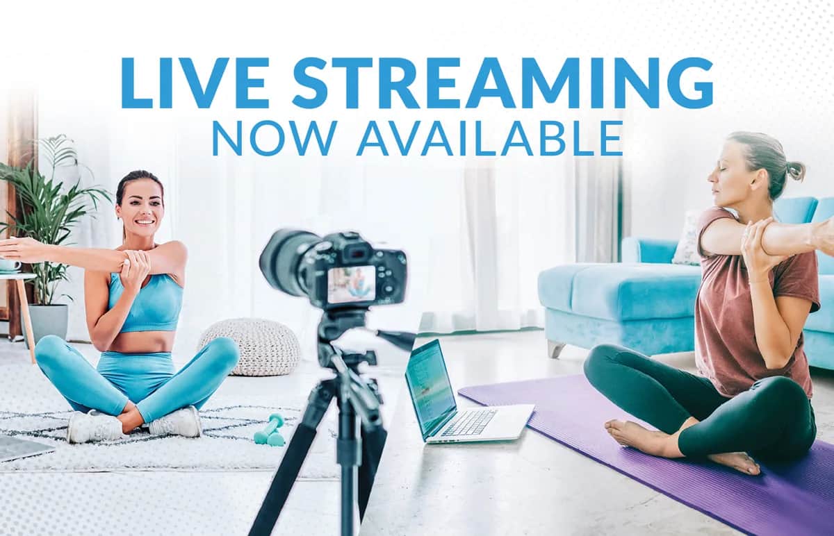 Two women in athletic wear stretch on yoga mats in their homes, with a camera and laptop between them. Text above reads: Live streaming now available.